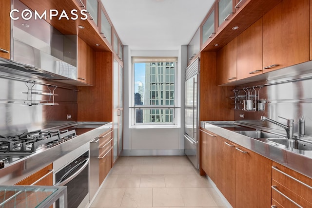 kitchen featuring stainless steel appliances, a sink, backsplash, glass insert cabinets, and brown cabinetry