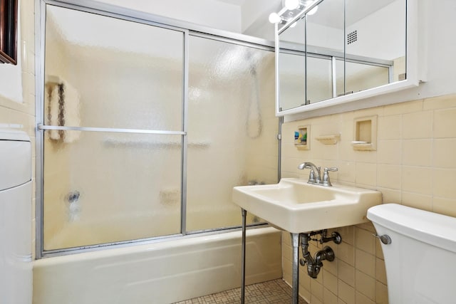 full bathroom featuring tile walls, toilet, bath / shower combo with glass door, washer / dryer, and a sink