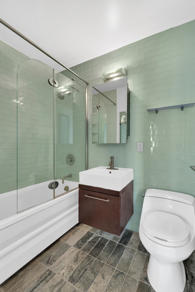 bathroom with vanity, toilet, shower / bath combination with glass door, tile walls, and stone finish flooring