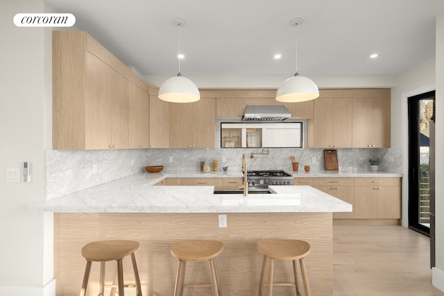 kitchen featuring light brown cabinets, a sink, backsplash, wall chimney exhaust hood, and a breakfast bar area