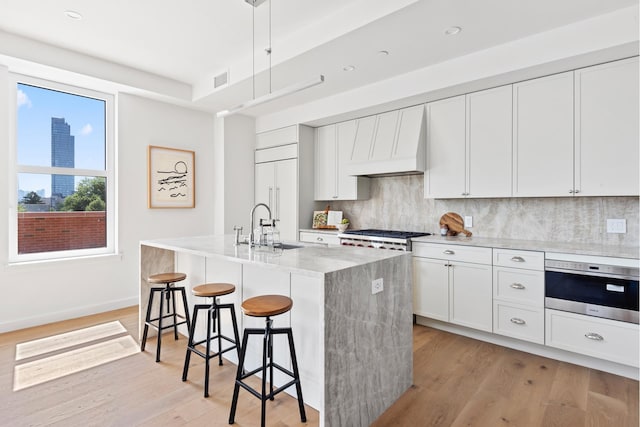 kitchen with custom exhaust hood, stainless steel oven, backsplash, light wood-type flooring, and a sink