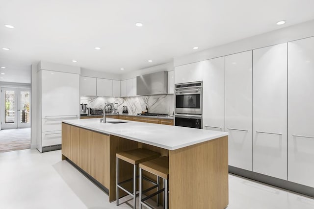 kitchen with a breakfast bar area, white cabinets, stainless steel appliances, a center island with sink, and wall chimney exhaust hood