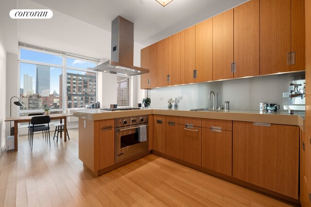 kitchen featuring oven, kitchen peninsula, sink, island exhaust hood, and light hardwood / wood-style flooring