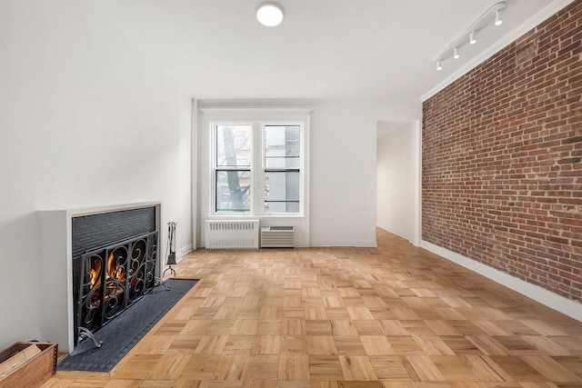 unfurnished living room featuring a warm lit fireplace, baseboards, radiator, brick wall, and track lighting