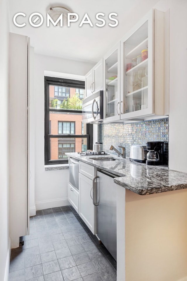 kitchen featuring glass insert cabinets, decorative backsplash, dark stone counters, stainless steel microwave, and baseboards