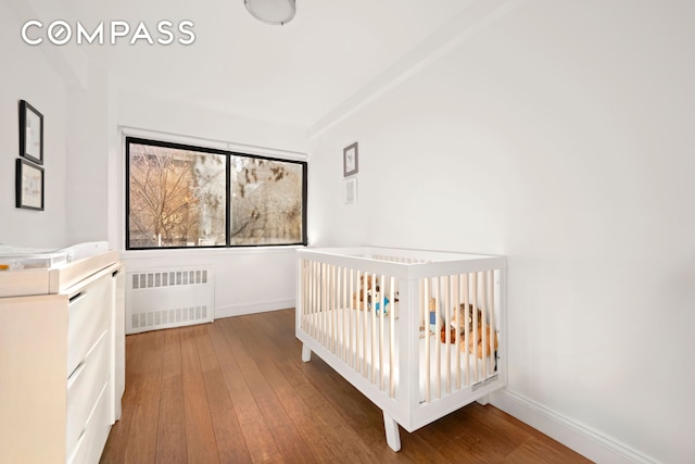 unfurnished bedroom with baseboards, a nursery area, wood-type flooring, and radiator