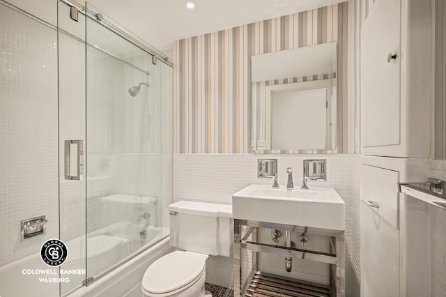 bathroom featuring toilet, a sink, shower / bath combination with glass door, tile walls, and wallpapered walls