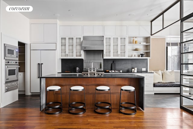 kitchen with white cabinetry, a kitchen island with sink, exhaust hood, and built in appliances