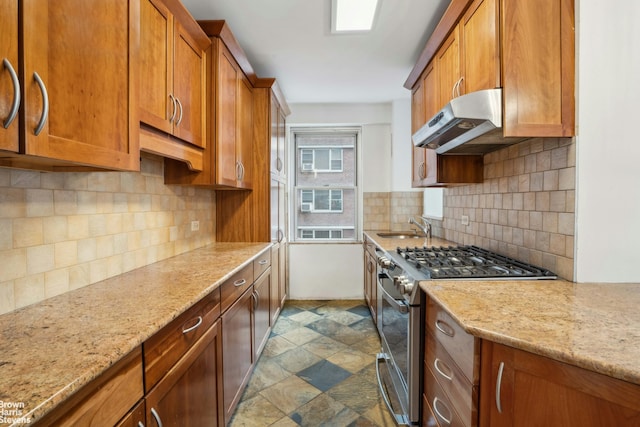 kitchen with stone finish floor, brown cabinets, stainless steel range with gas stovetop, under cabinet range hood, and a sink