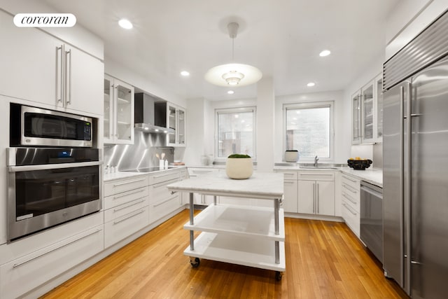 kitchen featuring appliances with stainless steel finishes, pendant lighting, white cabinets, wall chimney range hood, and light wood-type flooring