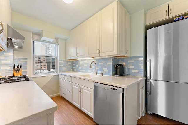 kitchen featuring wall chimney range hood, light countertops, a sink, light wood-style flooring, and stainless steel appliances