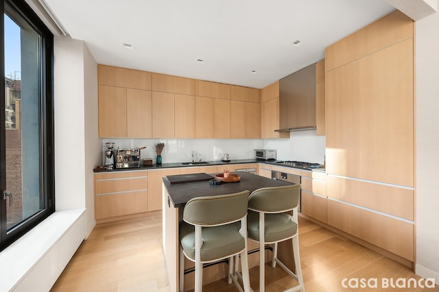 kitchen featuring light brown cabinets, wall chimney range hood, dark countertops, and a kitchen island