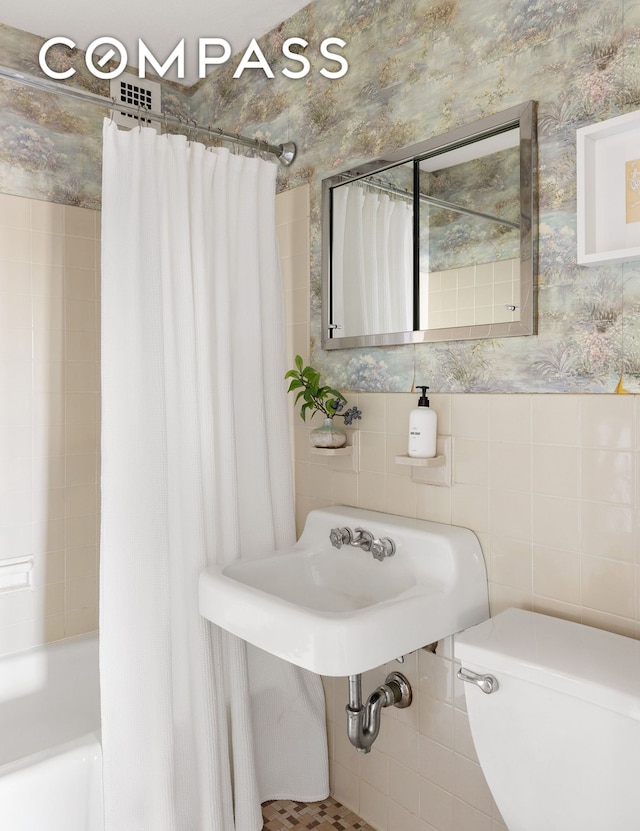 full bath with wallpapered walls, tile walls, toilet, shower / bathtub combination with curtain, and a sink