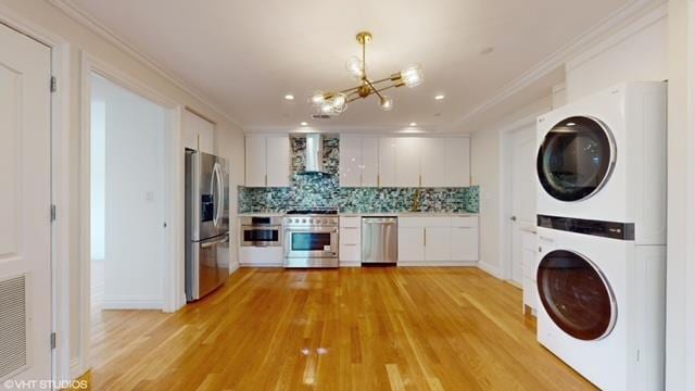 kitchen featuring stacked washer / dryer, appliances with stainless steel finishes, light countertops, wall chimney range hood, and white cabinetry