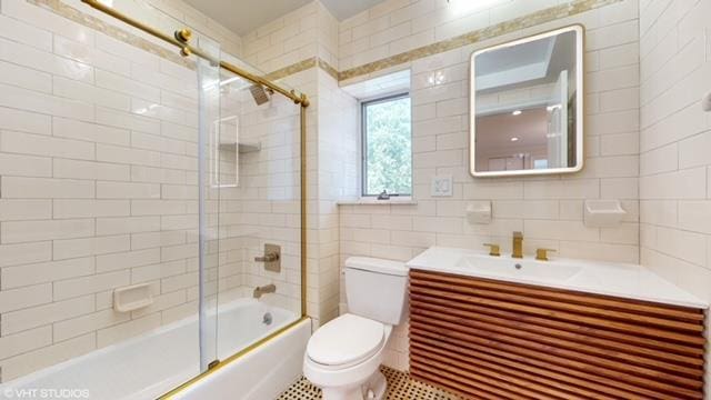 bathroom featuring tasteful backsplash, toilet, shower / bath combination with glass door, vanity, and tile walls