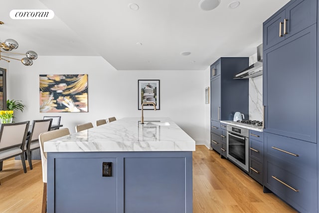 kitchen with sink, a center island with sink, light wood-type flooring, appliances with stainless steel finishes, and wall chimney range hood