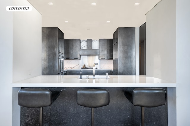 kitchen featuring a peninsula, wall chimney range hood, a kitchen bar, and light countertops