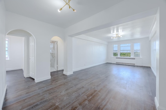 unfurnished living room featuring radiator, an inviting chandelier, wood finished floors, arched walkways, and baseboards