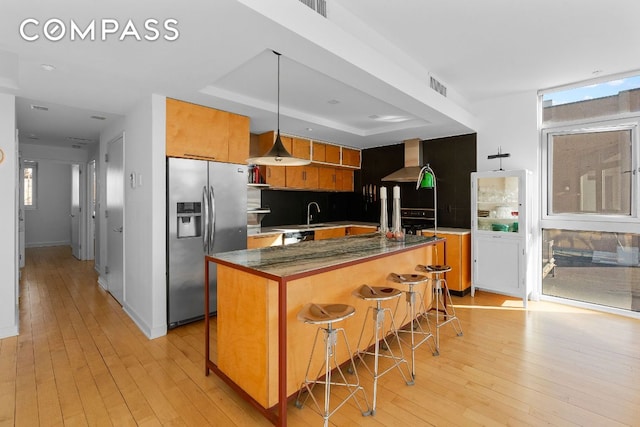 kitchen featuring stainless steel refrigerator with ice dispenser, light hardwood / wood-style floors, a kitchen bar, decorative light fixtures, and wall chimney exhaust hood