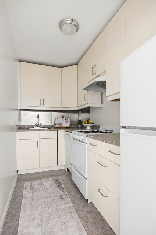 kitchen with white appliances, light tile patterned floors, cream cabinetry, under cabinet range hood, and a sink