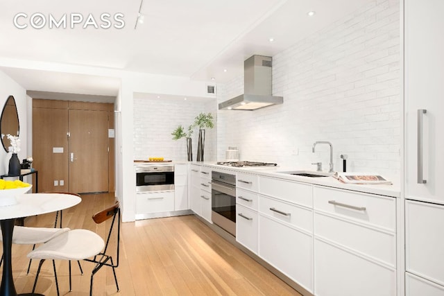 kitchen featuring white cabinetry, sink, wall chimney exhaust hood, stainless steel appliances, and light hardwood / wood-style flooring