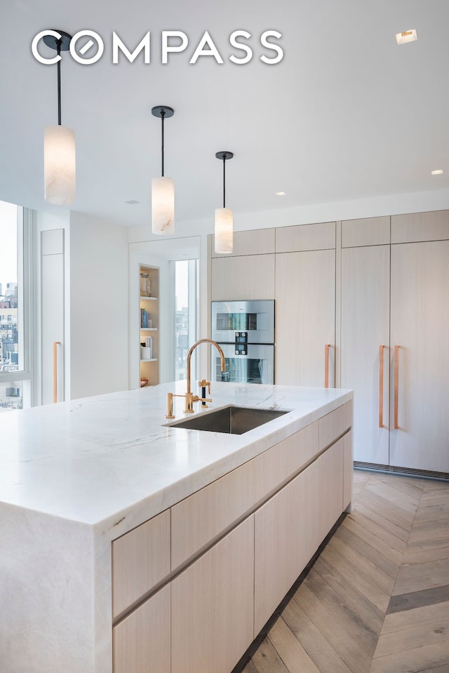 kitchen featuring light stone counters, pendant lighting, stainless steel double oven, a sink, and an island with sink
