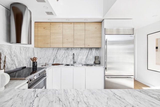 kitchen with light stone counters, light brown cabinetry, a sink, built in fridge, and range