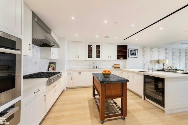 kitchen with appliances with stainless steel finishes, wall chimney exhaust hood, beverage cooler, sink, and white cabinetry