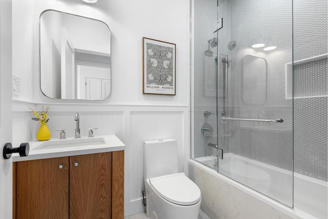 bathroom with shower / bath combination with glass door, wainscoting, vanity, and toilet