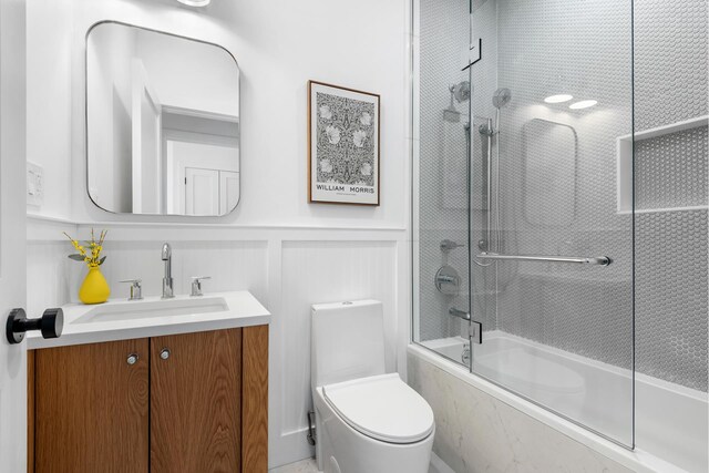 bathroom with toilet, vanity, combined bath / shower with glass door, a decorative wall, and wainscoting