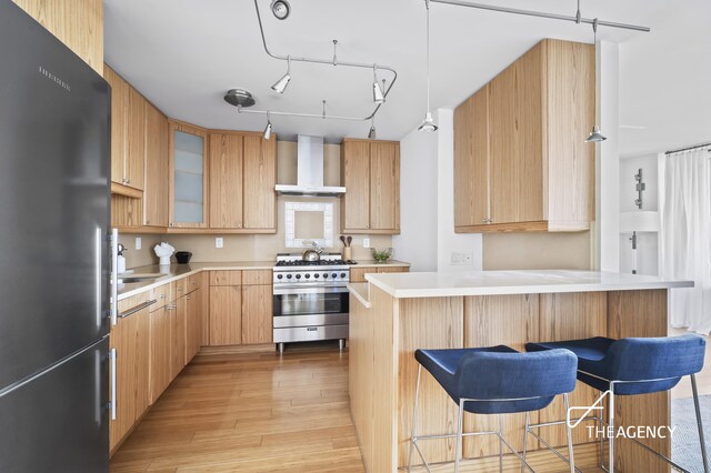 kitchen featuring light wood finished floors, wall chimney exhaust hood, high end stainless steel range, freestanding refrigerator, and a kitchen bar