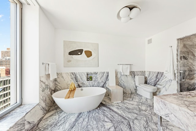 full bath featuring marble finish floor, tile walls, a soaking tub, toilet, and wainscoting