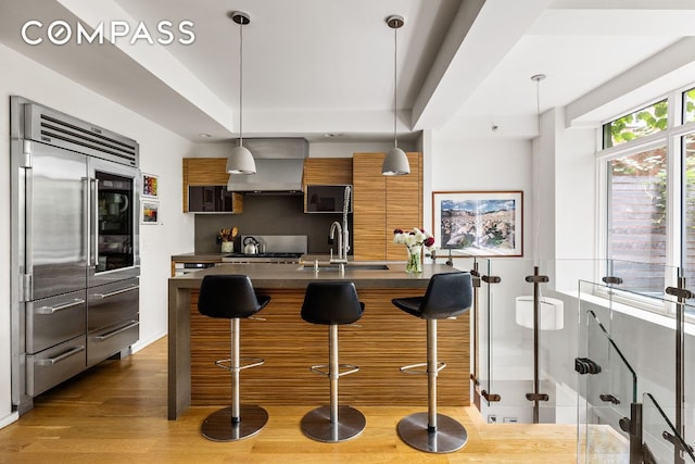 kitchen with stainless steel built in fridge, wall chimney exhaust hood, a kitchen bar, and decorative light fixtures