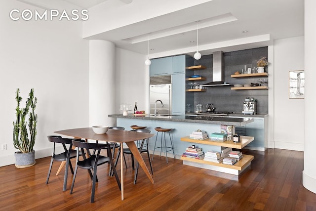 bar with sink, built in refrigerator, blue cabinets, decorative backsplash, and wall chimney exhaust hood