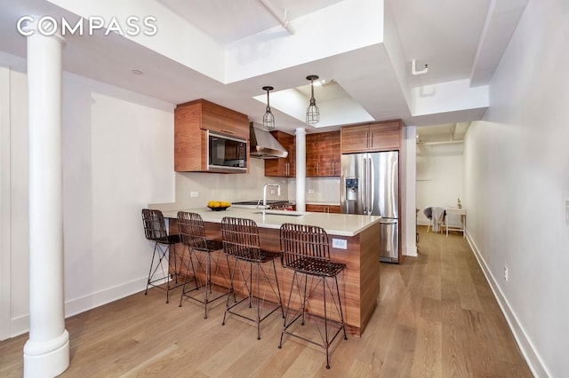 kitchen with a tray ceiling, stainless steel fridge with ice dispenser, brown cabinetry, built in microwave, and wall chimney exhaust hood