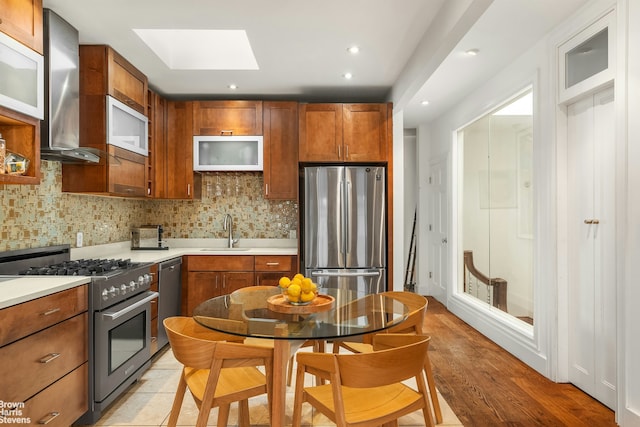 kitchen featuring a sink, appliances with stainless steel finishes, a skylight, wall chimney range hood, and decorative backsplash