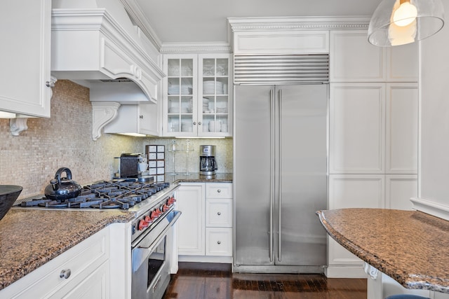 kitchen featuring premium appliances, tasteful backsplash, glass insert cabinets, white cabinetry, and dark stone countertops