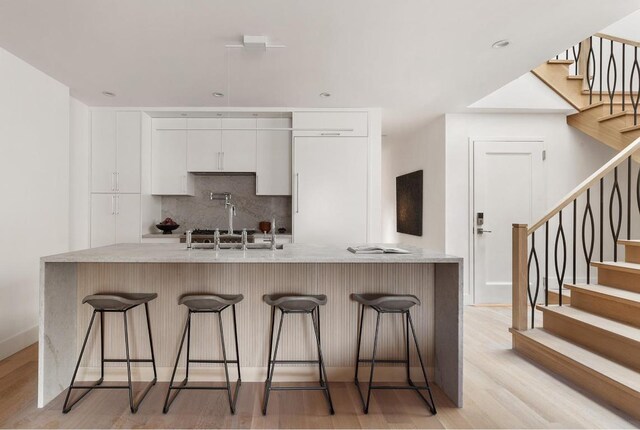 kitchen with a breakfast bar area, a kitchen island with sink, white cabinets, decorative backsplash, and light wood-type flooring