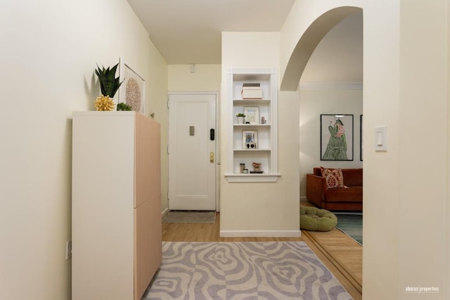 corridor featuring arched walkways, built in shelves, light wood-style flooring, and baseboards