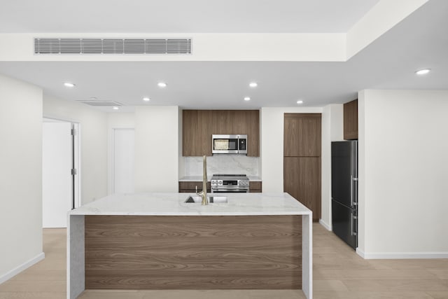 kitchen with light wood-type flooring, a center island with sink, stainless steel appliances, light stone countertops, and tasteful backsplash