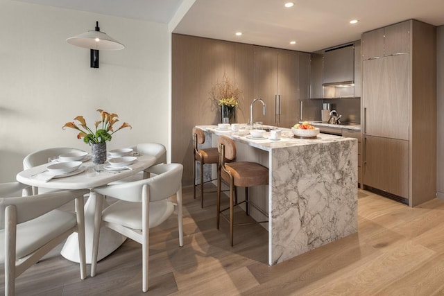 kitchen featuring light wood-type flooring, black electric cooktop, a kitchen breakfast bar, an island with sink, and light stone countertops