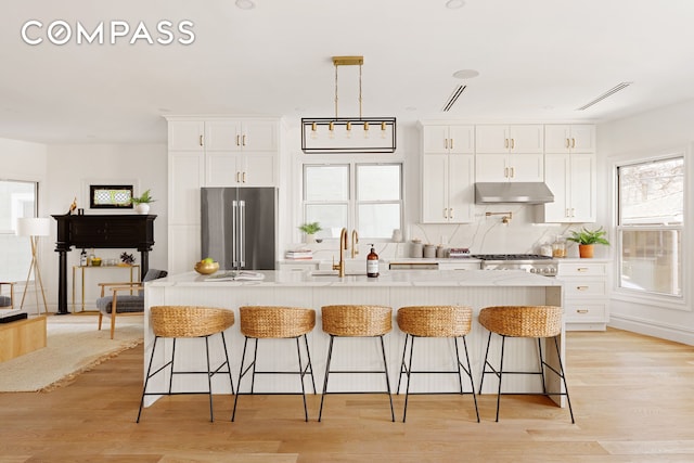 kitchen featuring white cabinets, a sink, high end refrigerator, under cabinet range hood, and a center island with sink