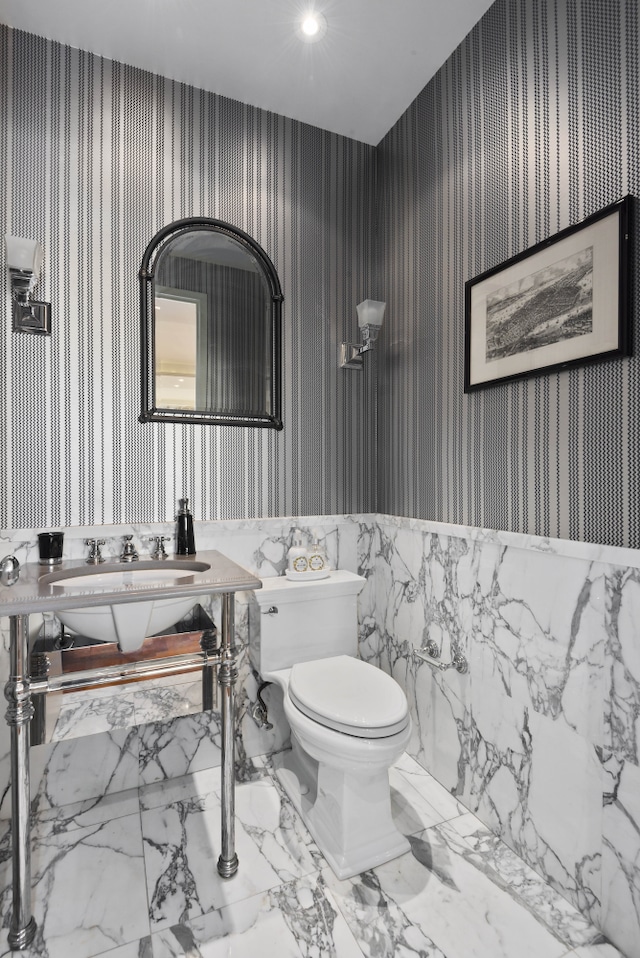 half bathroom with marble finish floor, a sink, toilet, and wallpapered walls