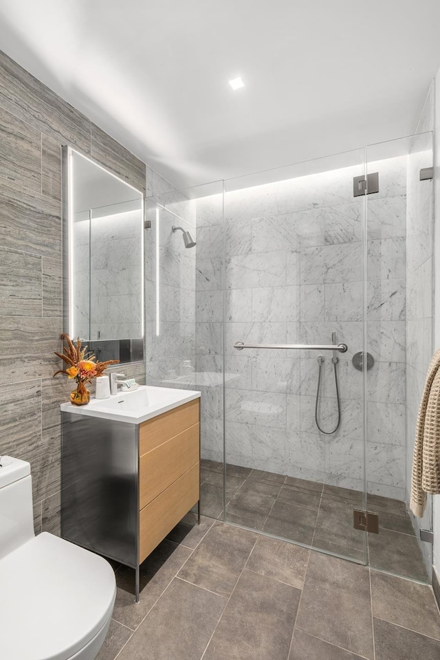 bathroom featuring toilet, a marble finish shower, tile walls, and vanity