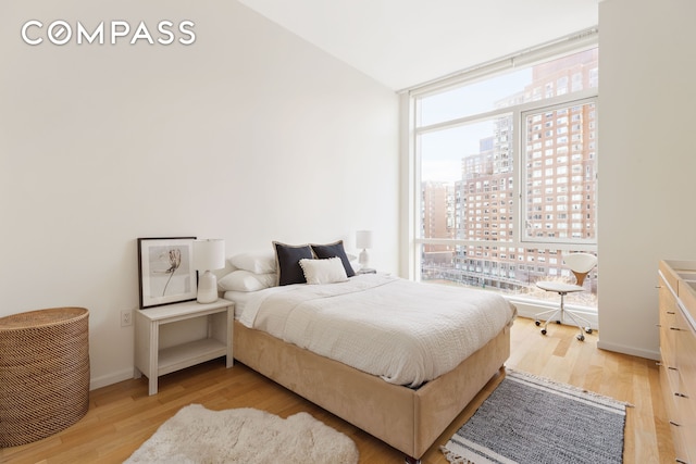 bedroom featuring a city view, light wood finished floors, vaulted ceiling, a wall of windows, and baseboards