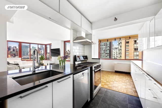 kitchen with wall chimney range hood, appliances with stainless steel finishes, white cabinetry, and a sink