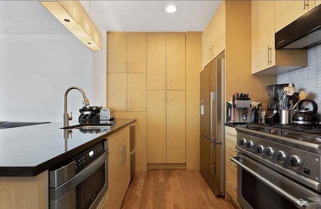 kitchen with dark countertops, high quality appliances, under cabinet range hood, and light brown cabinetry