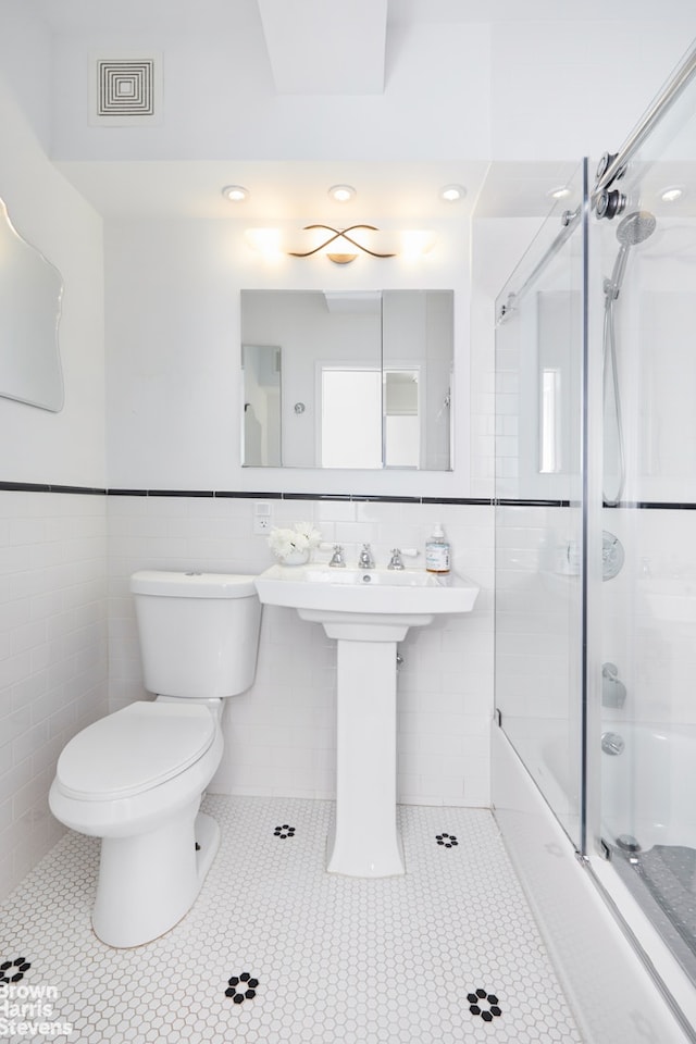 bathroom featuring tile walls, toilet, combined bath / shower with glass door, tile patterned floors, and a wainscoted wall
