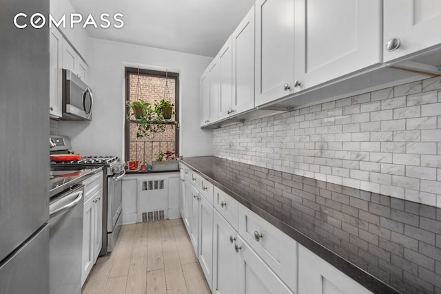 kitchen featuring light wood finished floors, tasteful backsplash, radiator heating unit, stainless steel appliances, and white cabinetry