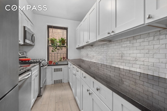 kitchen with tasteful backsplash, dark countertops, radiator heating unit, light wood-type flooring, and appliances with stainless steel finishes